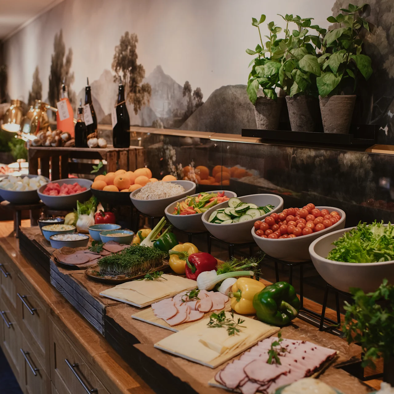 Et buffetbord med forskellige friske grøntsager, frugter, skiveskåret kød og urter arrangeret på en lang træbænk mod en vægmaleri af et naturlandskab.