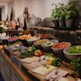 Ett buffébord med olika färska grönsaker, frukter, skivat kött och örter arrangerade på en lång träbänk mot en väggmålning som visar ett naturlandskap.