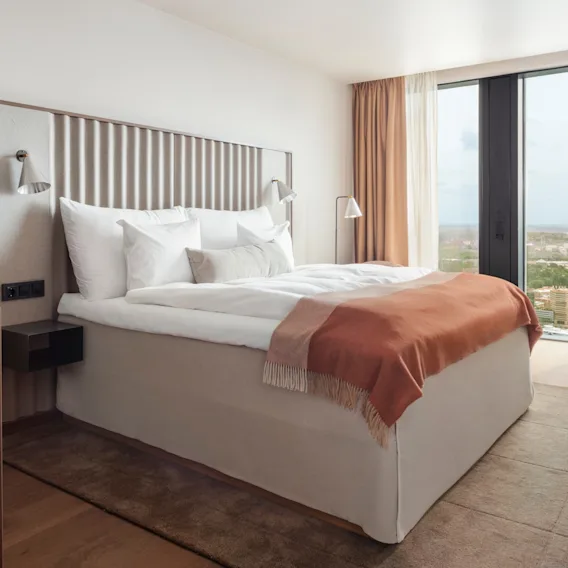 En pent seng med hvitt sengetøy og et oransje pledd i et moderne hotellrom. Store vinduer gir utsikt over landskapet utenfor.