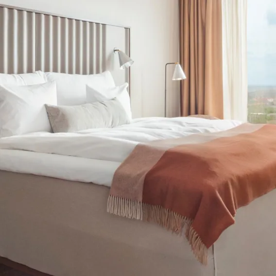 En snyggt bäddad säng med vita sängkläder och en orange pläd i ett modernt hotellrum. Stora fönster ger utsikt över landskapet utanför.