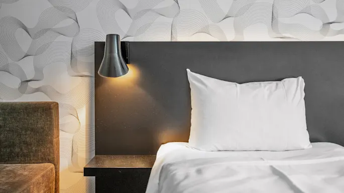 White pillow on a bed with an illuminated wall-mounted lamp; background has a wavy patterned wallpaper.