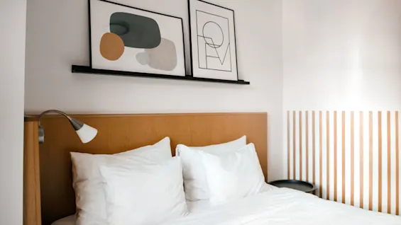 A neatly made bed with white bedding, wooden headboard, abstract art above, and a small lamp mounted to the wall in a minimalist room.