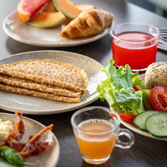 A breakfast spread featuring flatbread, salad with tomatoes and cucumbers, and juices. Surrounding the plates are croissants, melon slices, and scrambled eggs on a dark wooden table.