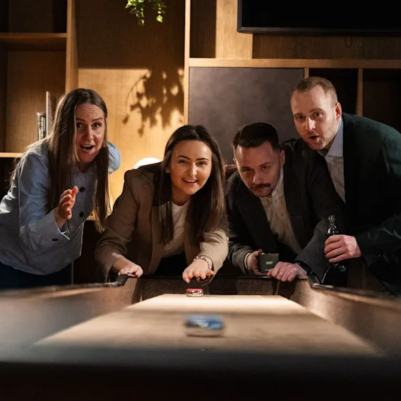 Fire engasjerte kollegaer som følger spent med mens de spiller shuffleboard sammen i loungen.