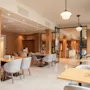En restaurant med beige stoler og trebord viser et tomt sitteområde. Moderne lysarmaturer henger fra taket. Glassvegger deler opp rom, og skaper en innbydende atmosfære.