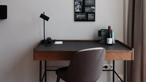 Ett modernt skrivbord med lampa, telefon och papper i ett rum med affischer, en vinflaska och en stol.