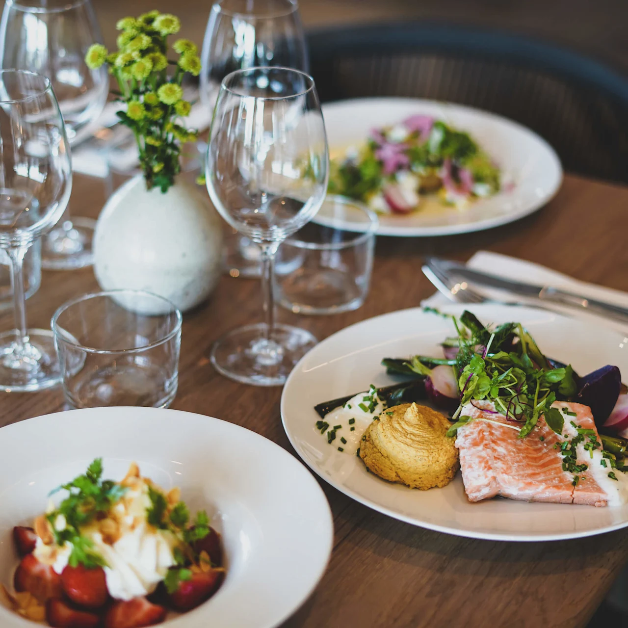 Tallerkener med gourmetmat er elegant satt opp på et trebord, omgitt av tomme vinglass og en liten vase med blomster, og skaper en raffinert middagssetting.