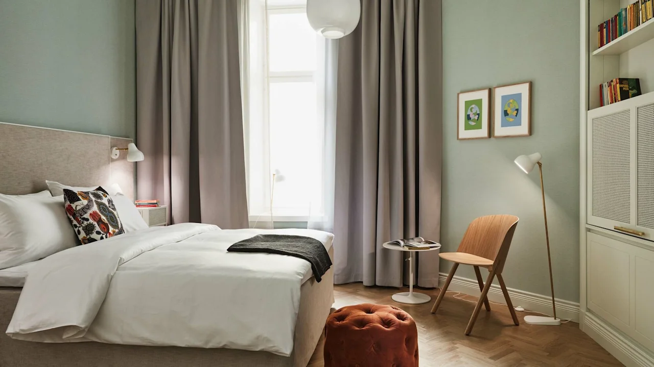A bright and modern hotel room featuring a made double bed, wooden chair, and soft green walls.