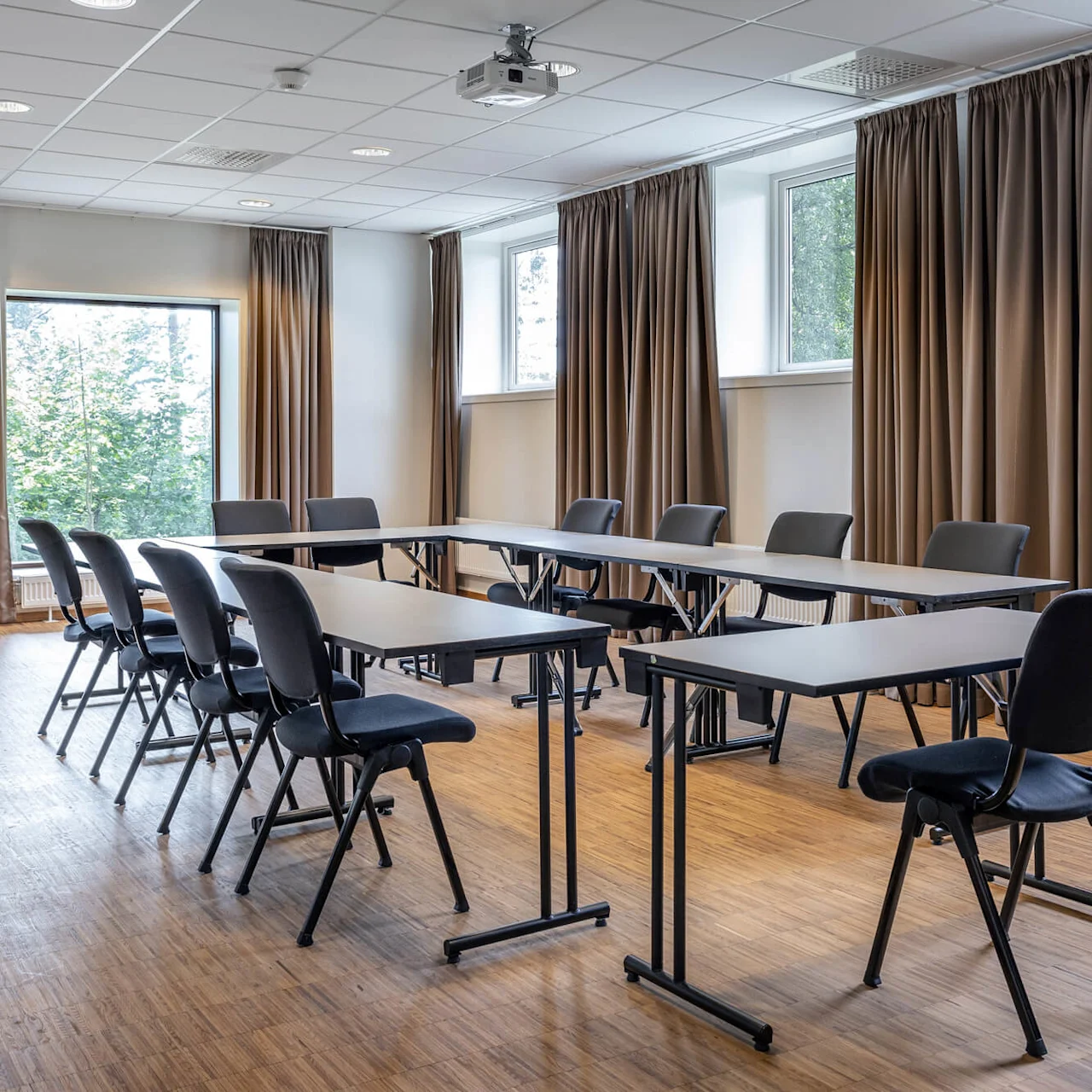 Konferansebord arrangert i en U-form med stoler rundt. Rommet har tregulv, taklamper og vinduer med brune gardiner, og skaper et profesjonelt møtemiljø.