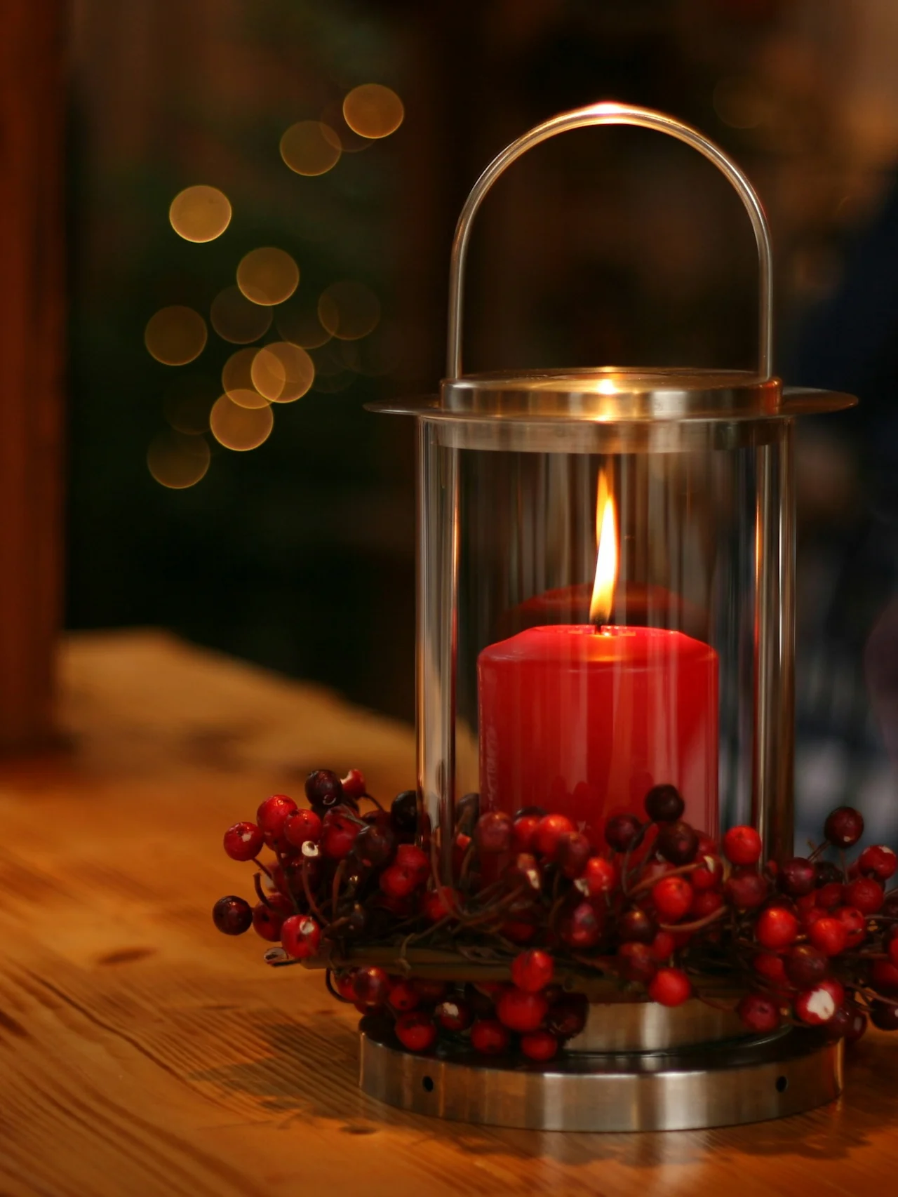 Ett rött ljus brinner i en glaslykta, dekorerad med röda bär, står på ett träbord. I bakgrunden antyder suddiga lampor en mysig inomhusmiljö.
