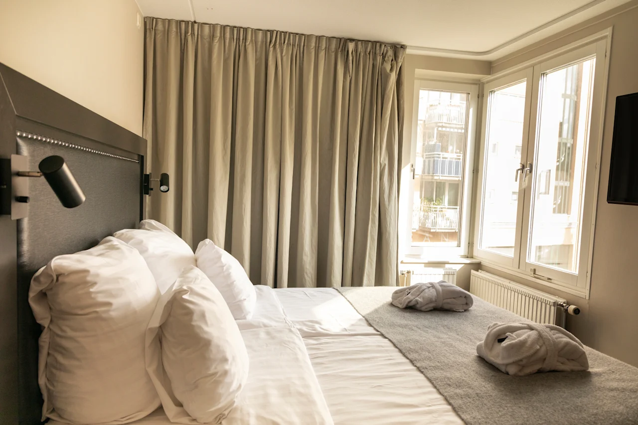 Ett ljust och inbjudande hotellrum med en bekväm säng med vita lakan, två badrockar och ett stort fönster.