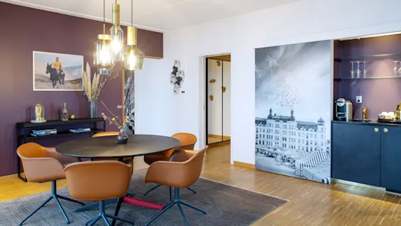 Ett runt matbord med bruna stolar står på en matta; hängande lampor belyser det moderna rummet dekorerat med väggkonst och ett stadsvykort. Skåp och dekorationer förstärker miljön.