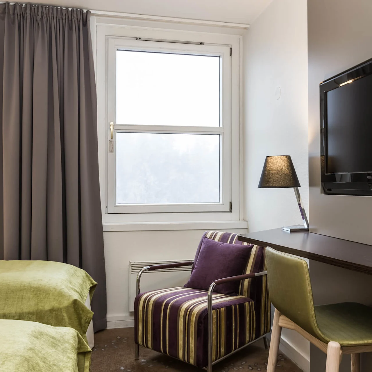 A wall-mounted TV is opposite a striped armchair near a window. A desk with a lamp and chair are beside two beds with green covers.