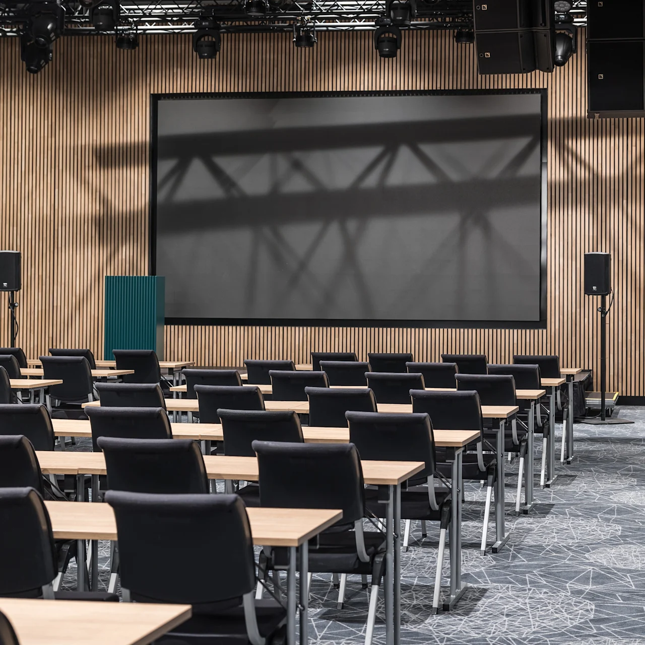 Konferenssihuone, jossa riveittäin tyhjiä tuoleja ja pöytiä, suuntautuu kohti suurta, tyhjää projektoripintaa puupaneeliseinällä, ympäröity kaiuttimilla ja valaisimilla.