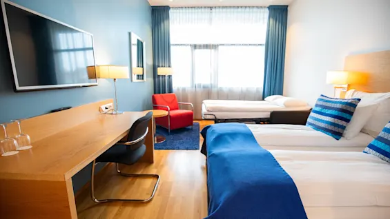 Et ryddig hotellrom har en dobbeltseng med blå detaljer, et skrivebord i tre og en veggmontert TV. En rød lenestol står ved et stort vindu med blå gardiner.