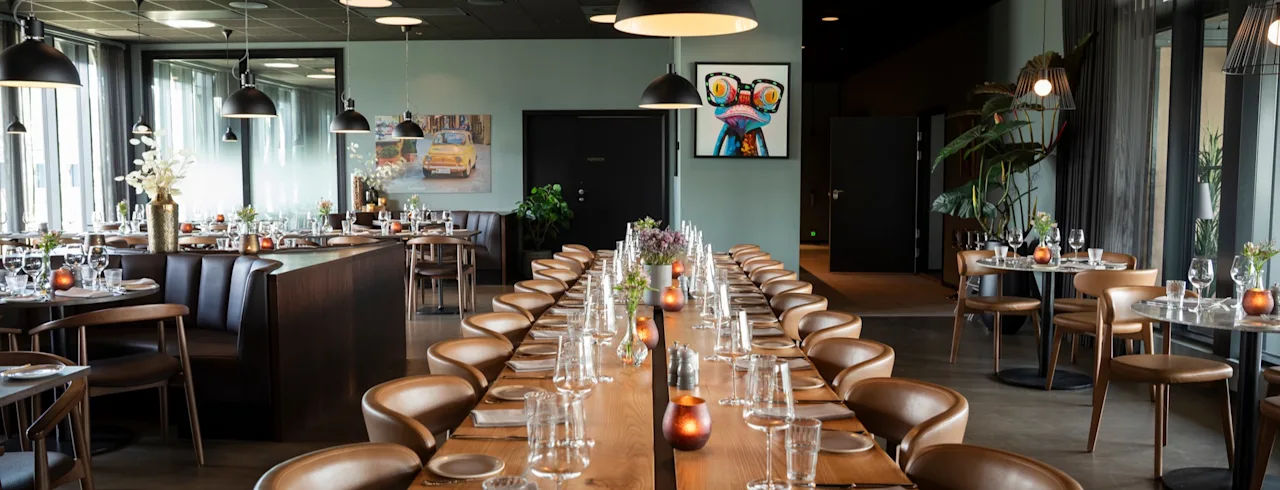Et moderne restaurantinteriør med et langt spisebord dækket til en stor forsamling, omgivet af mindre borde.