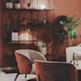 Two cushioned chairs sit on a circular rug, facing a metal shelf with decorative items, plants, and a lamp. A large plant adds greenery to the warm, ambient room.