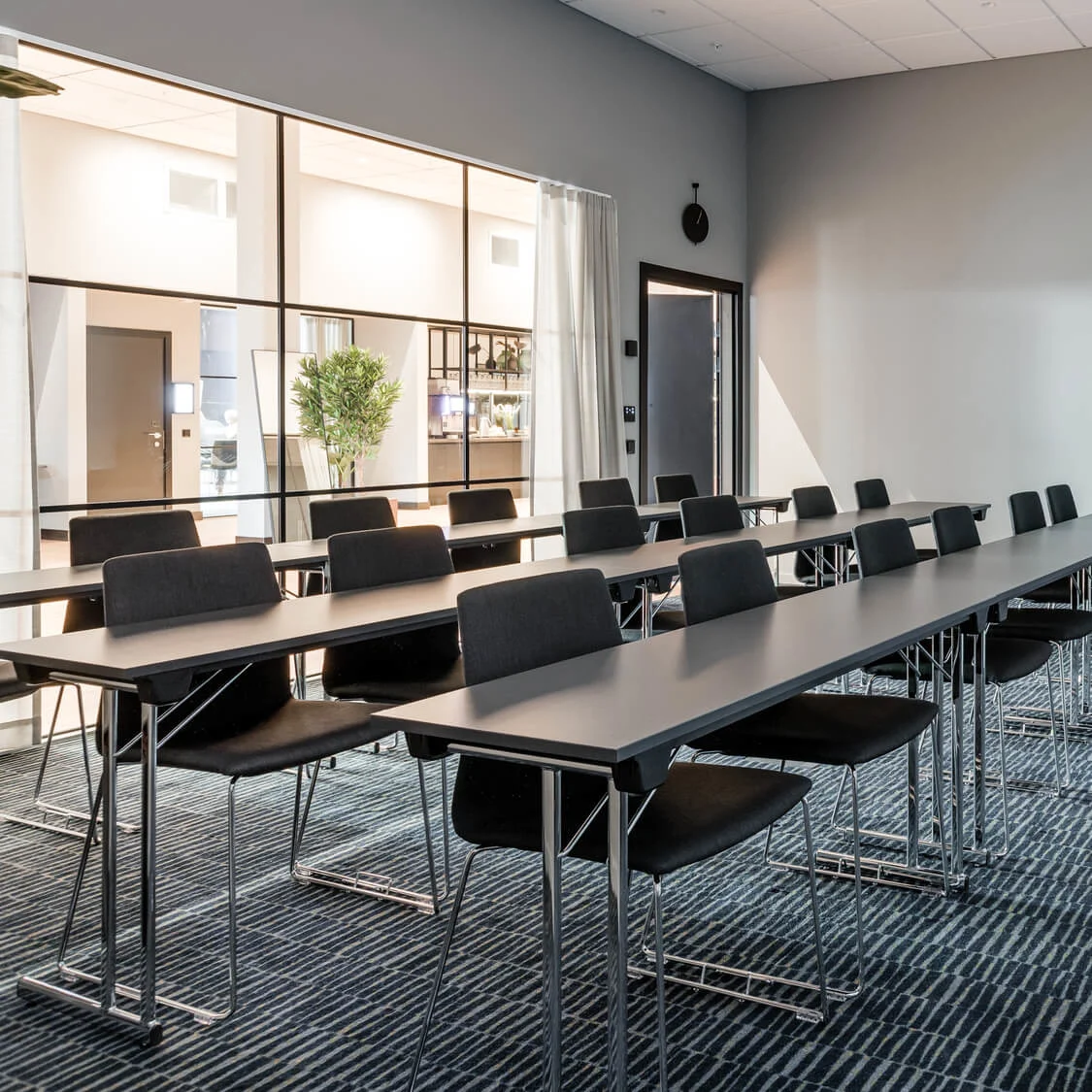 Rækker af tomme borde og stole er opstillet i et moderne konferencelokale. Et stort vindue adskiller lokalet fra et lyst interiørrum med planter.