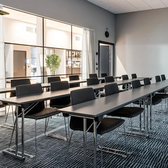 Rows of empty tables and chairs are lined up in a modern conference room. A large window separates the room from a bright interior space with plants.