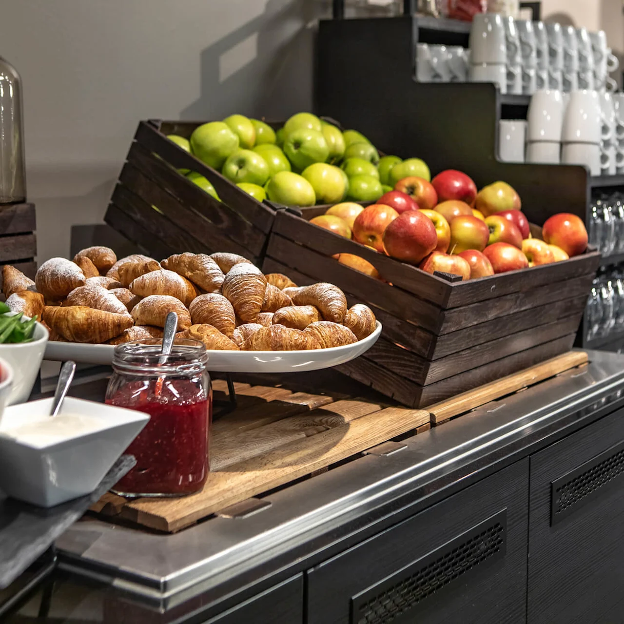 Croissants rest on a white plate beside jars of red jam on a buffet table, with crates of apples and green grapes in the background.