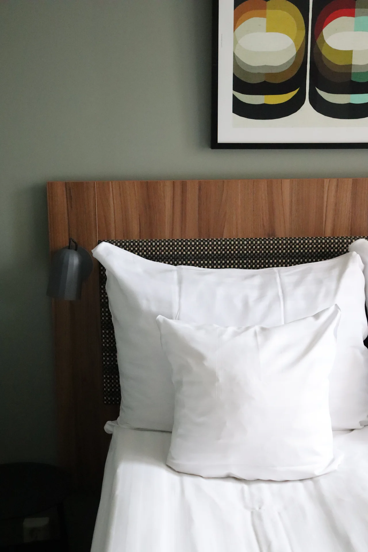 White pillows rest against a wooden headboard in a neatly made bed, beside a black lamp. Above, a colorful abstract artwork decorates the gray wall.