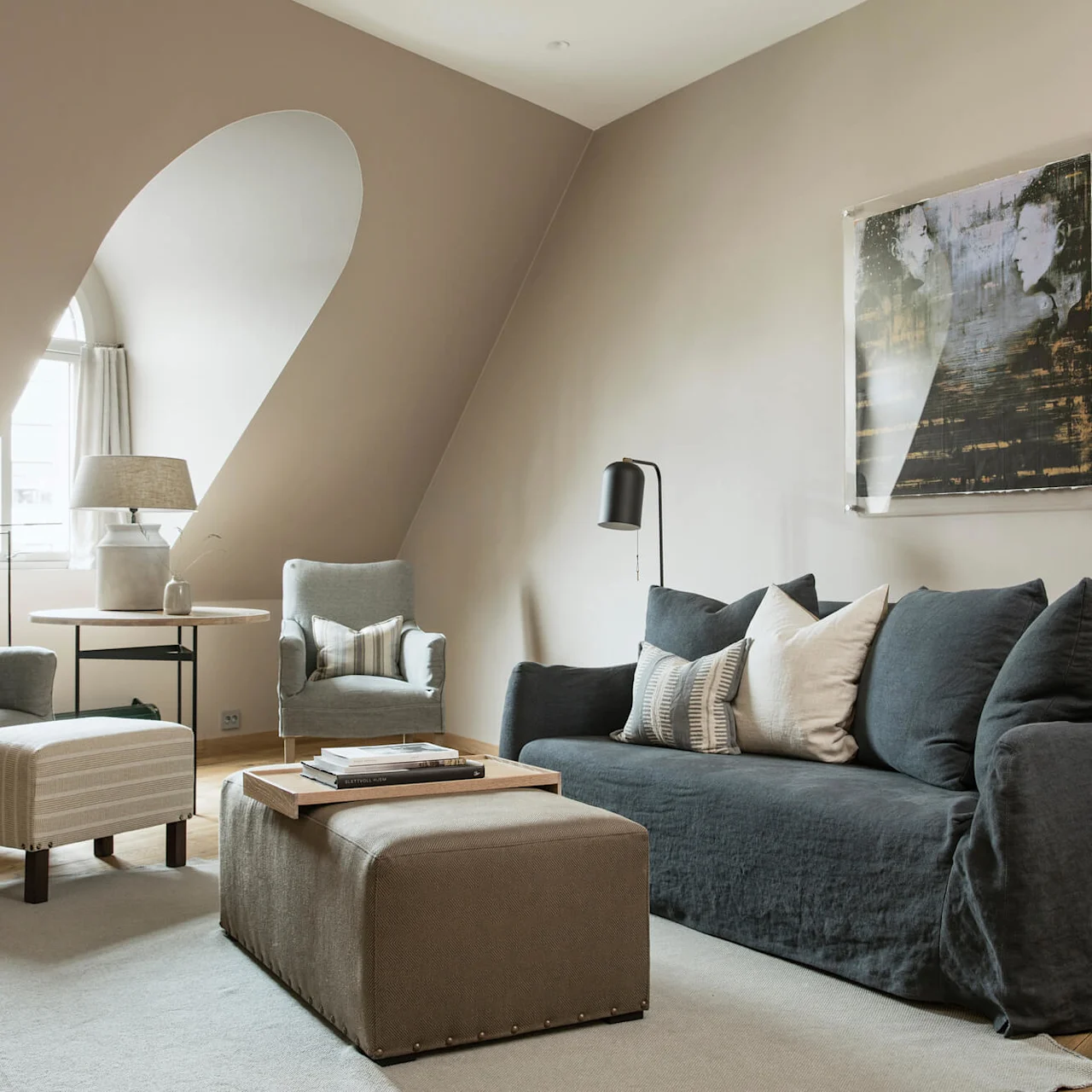 A cozy living room features a dark sofa with pillows, two armchairs, and a small table by a window. A framed artwork hangs on a sloped beige wall.