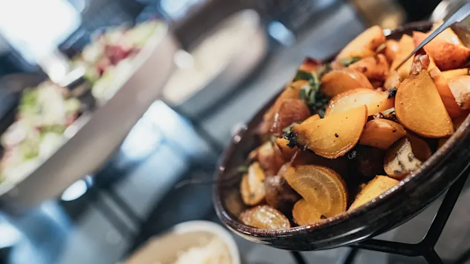 Roasted potatoes and herbs in a bowl, focused in foreground, with blurred restaurant setting behind.