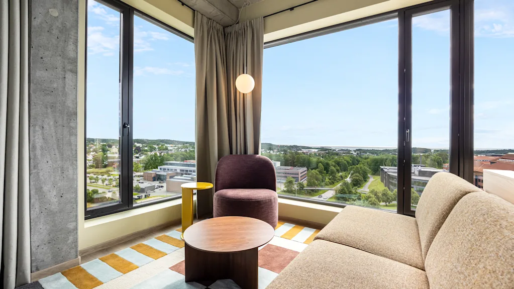 A modern hotel room with a comfortable sofa and armchair, featuring large windows that offer a panoramic view of the city.
