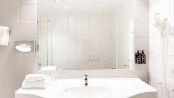 Et lyst, rent baderom med hvite fliser, et stort speil over en vask, friske håndklær og toalettartikler.