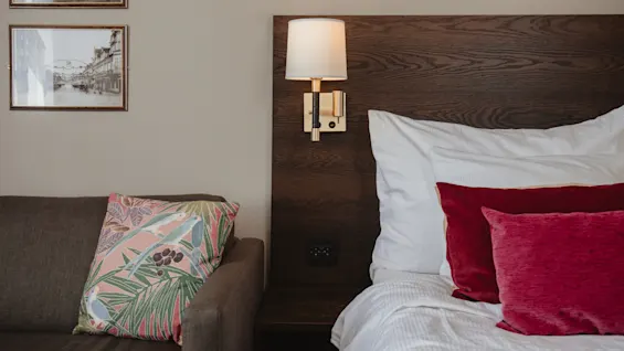 Seng med røde og hvide puder står mod et træpanel. En tændt væglampe er fastgjort over hovedgavlen. I nærheden står en brun sofa med en farverig blomsterpude.