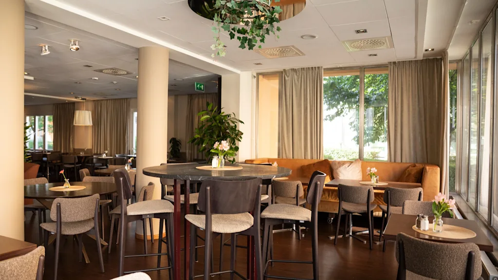En modern och inbjudande hotellounge eller restaurang med olika bord och sittplatser, inklusive en lång soffa vid stora fönster. EXIT-skylt synlig.