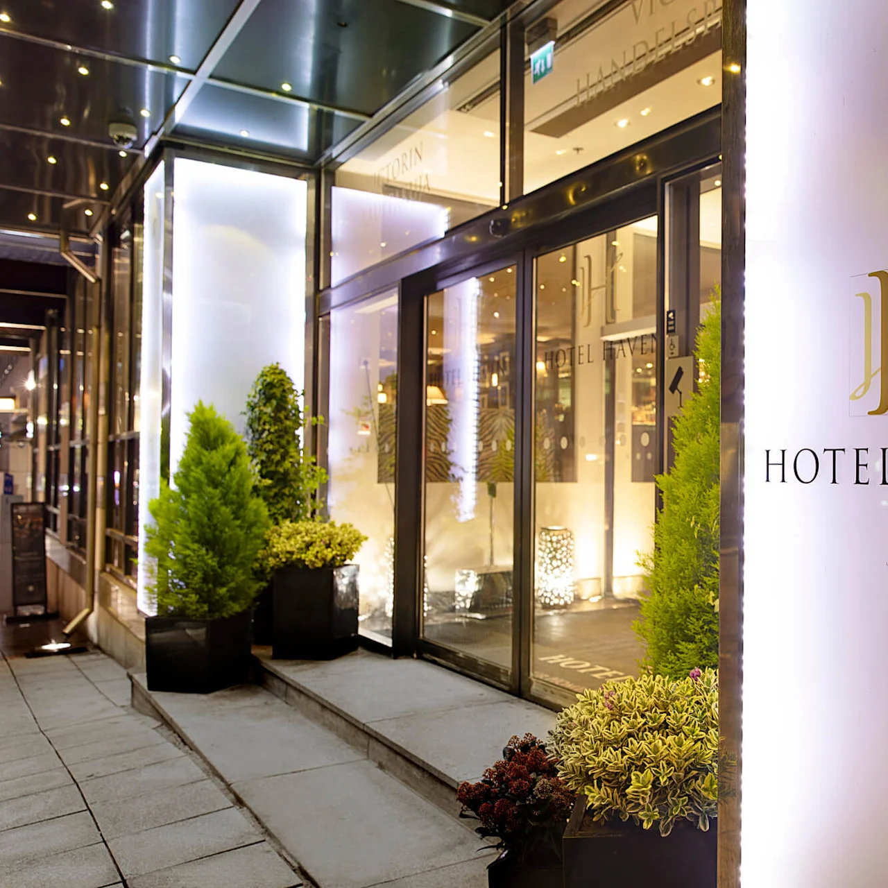 A hotel entrance features a glass door surrounded by potted plants. The sign reads "HOTEL HAVEN" in elegant lettering. The setting is illuminated and inviting.