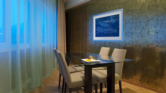A dining room corner with a glass table, four patterned chairs, and a picture of fish on a textured wall, illuminated by wall sconces.