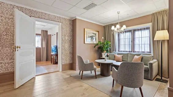 A cozy and elegantly furnished living area with a sofa, armchairs, and a coffee table, leading to another room through an open door.