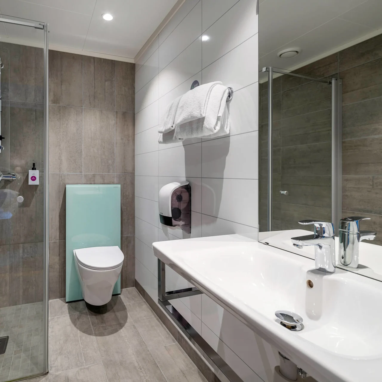 Shower stall stands enclosed by glass in a modern bathroom with toilet, sink, and towels on a rack. Neutral tiled walls and floor with a large mirror create a sleek ambiance. No text present.