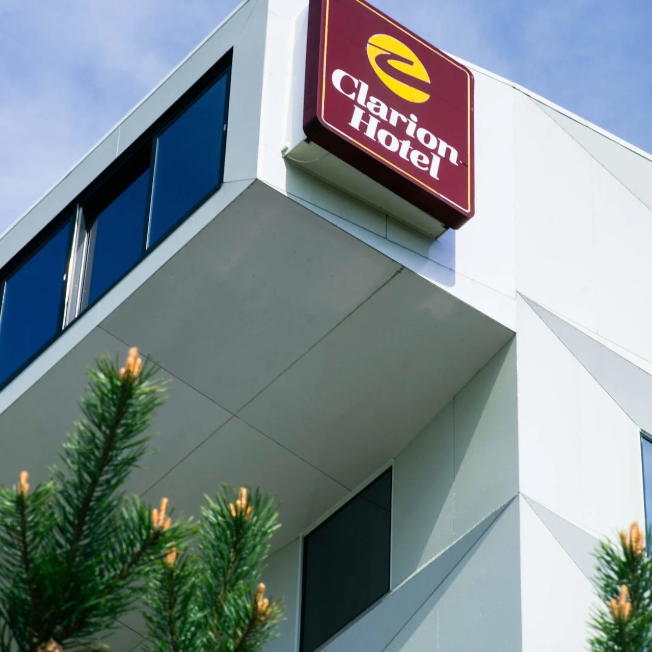 Et rødt skilt med gult emblem sier "Clarion Hotel" på hjørnet av en moderne hvit bygning, omgitt av blå himmel og grønne furugreiner.
