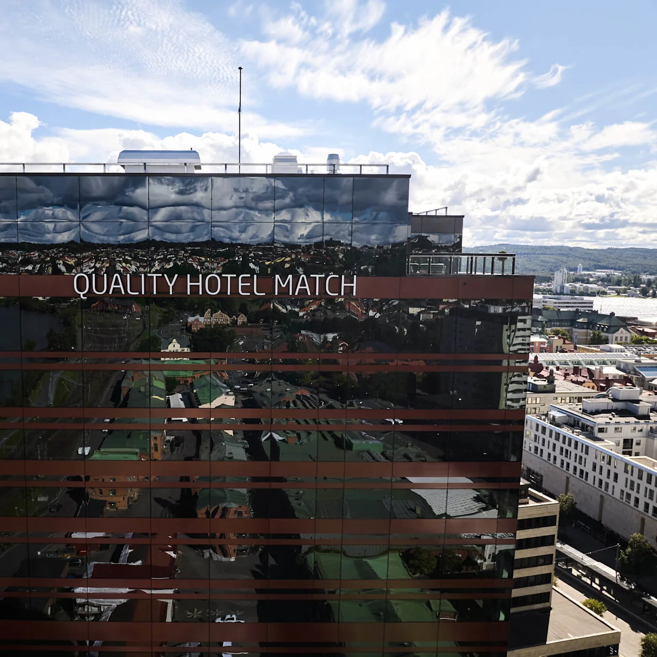 En reflekterende bygningsfacade viser "QUALITY HOTEL MATCH" i et bymiljø, med en naturskøn baggrund af vand, træer og byinfrastruktur under en delvis skyet himmel.