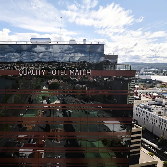 En reflekterende bygningfasade viser "QUALITY HOTEL MATCH" i et urbant miljø, med en naturskjønn bakgrunn av vann, trær og byinfrastruktur under en delvis skyet himmel.