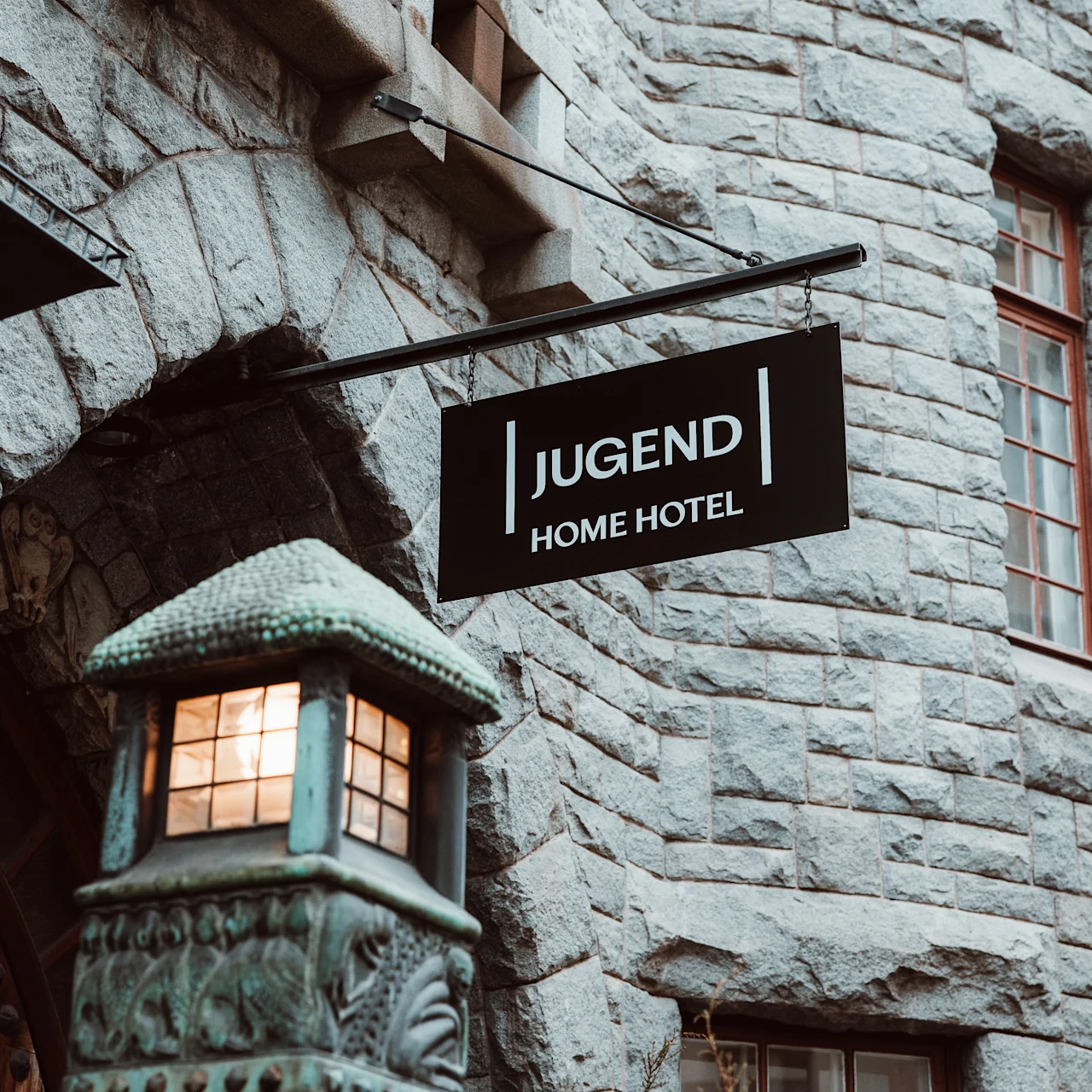 Et sort skilt med teksten "JUGEND HOME HOTEL" hænger fra en stenbygning. Nedenunder udsender en udsmykket gadelygte et varmt lys, hvilket fremhæver den historiske arkitektur.