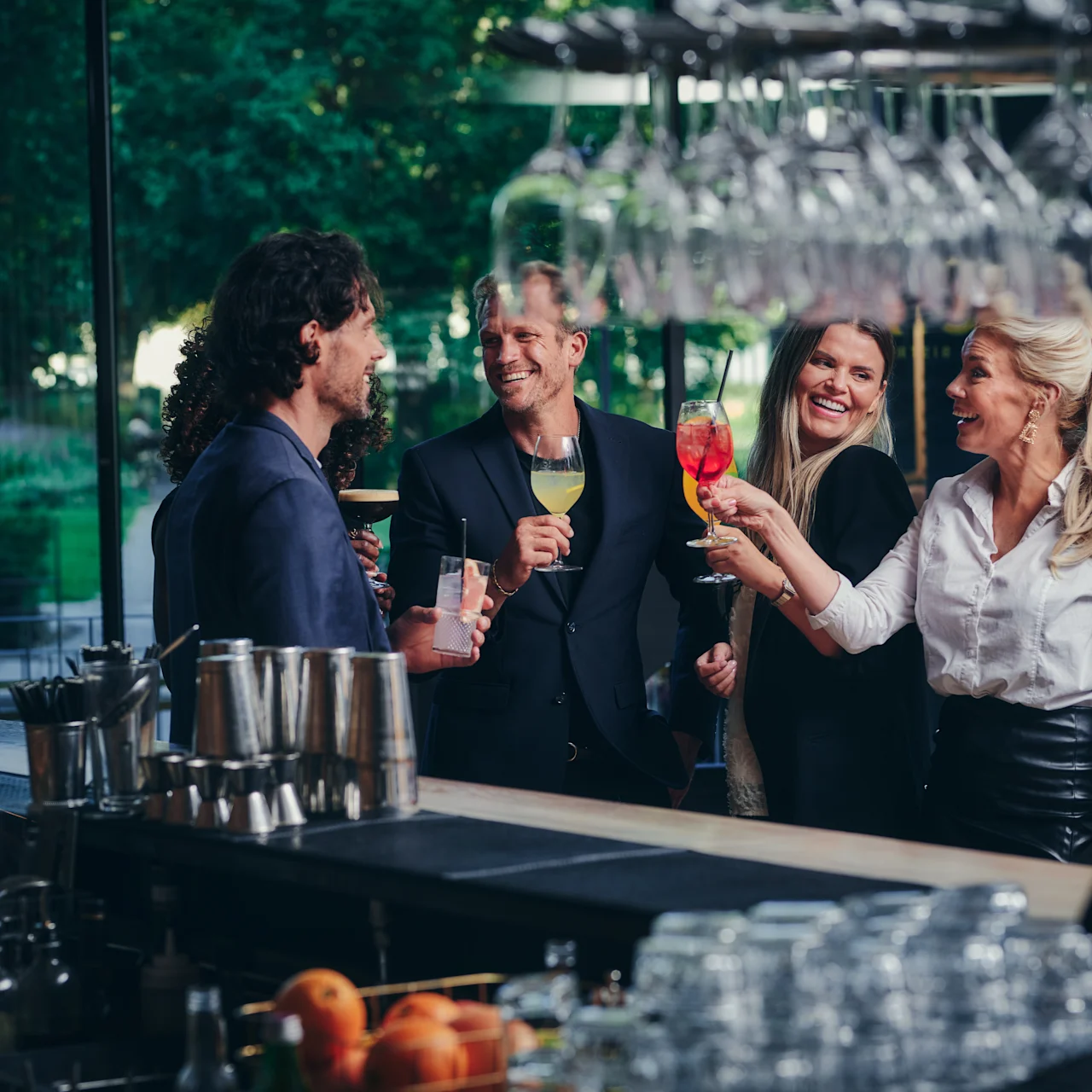 Fyra personer höjer glas, ler och står tillsammans vid en bar. Glas och frukt syns på disken, med träd som syns genom stora fönster.