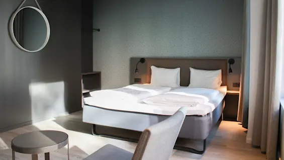 A neatly made double bed with white linens, flanked by wall lights, faces a round mirror. The room features minimalistic decor with gray walls and wooden flooring.