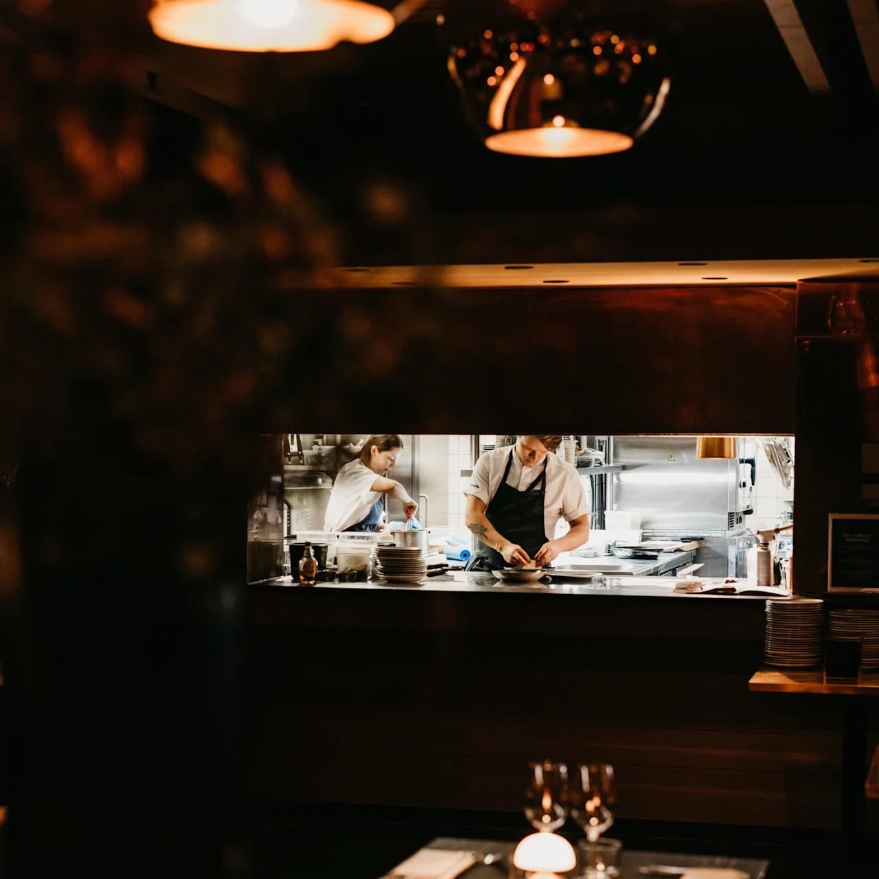 Chefs prepare food in a warmly lit restaurant kitchen viewed through a framed window.