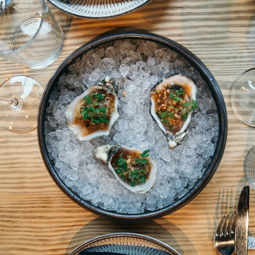 Tre garnerede østers på is i en skål, omgivet af vinglas og bestik på et træbord, klar til et måltid.