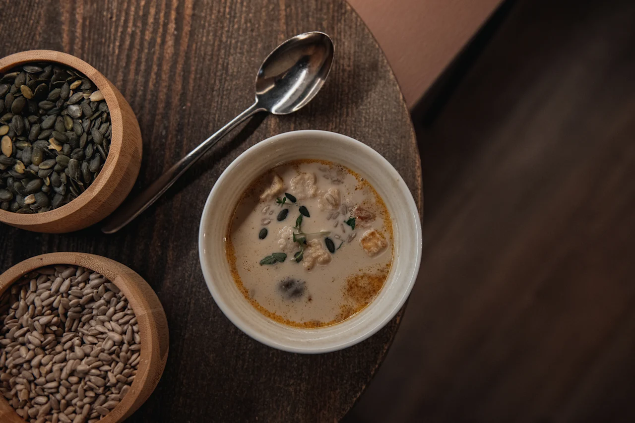 En varm skål krämig soppa med örter och frön, tillsammans med skålar med pumpa- och solrosfrön, på ett rustikt träbord.