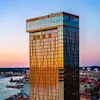 En hög glasbyggnad märkt "Hotel Draken" speglar en solnedgångssky över ett stadslandskap vid vattnet. I närheten rinner en flod bredvid urbana byggnader och kajer.