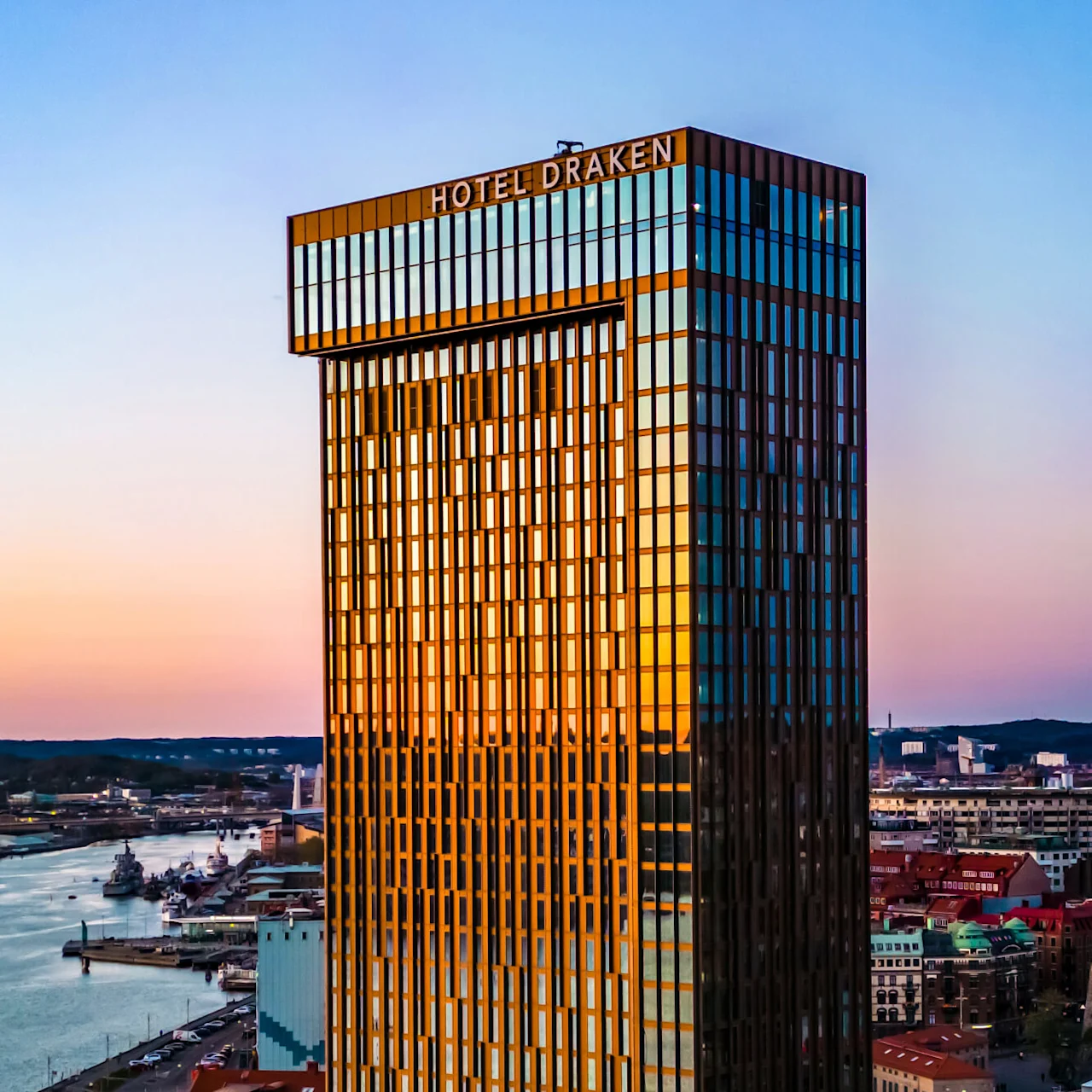 En hög glasbyggnad märkt "Hotel Draken" speglar en solnedgångssky över ett stadslandskap vid vattnet. I närheten rinner en flod bredvid urbana byggnader och kajer.