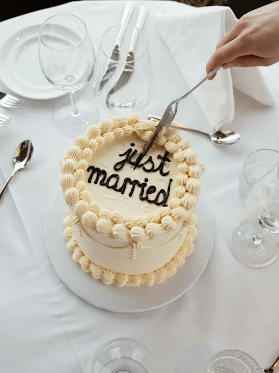 En hånd skjærer i en hvit kake med teksten 'just married', omgitt av dekket bord for en feiring.