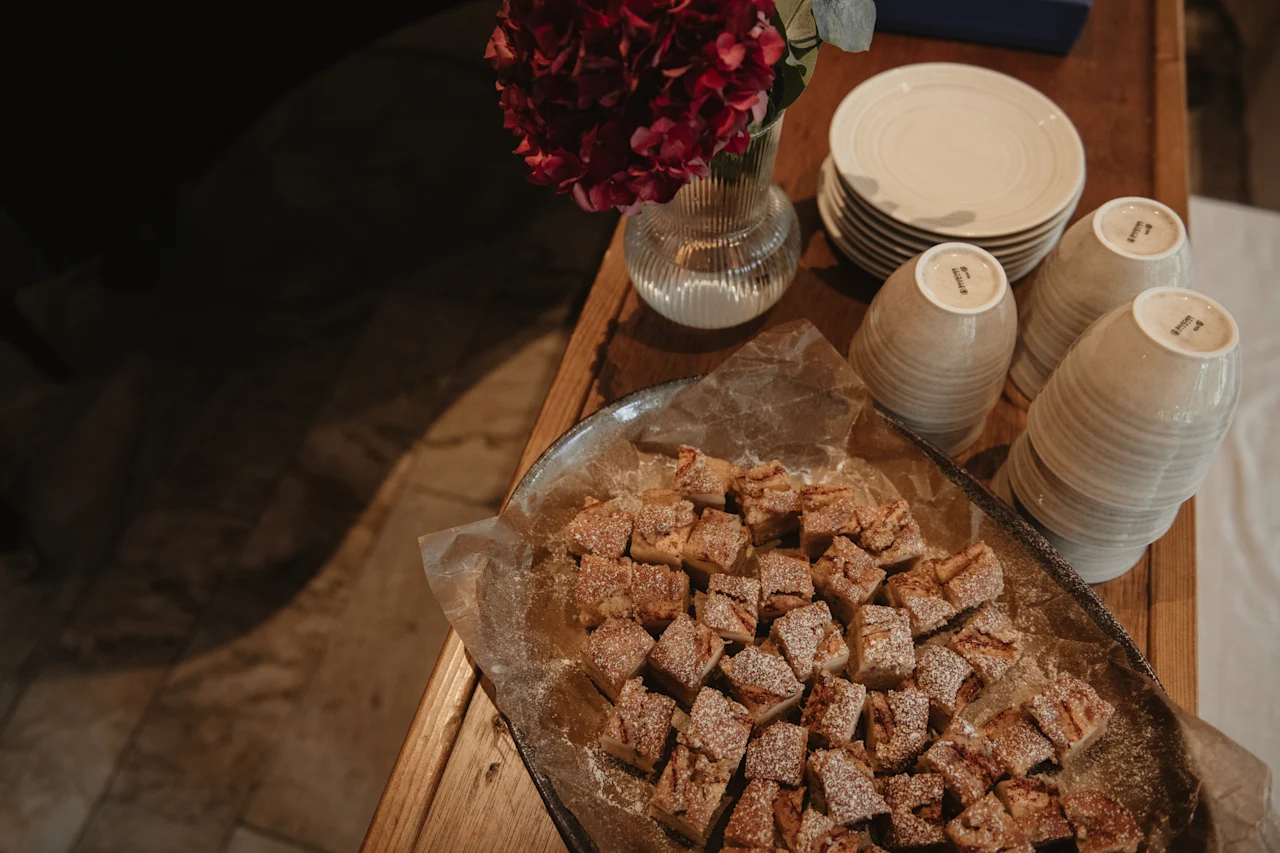 A table laden with delicious-looking pastries, a vase of red flowers, and stacks of plates and cups, ready for a gathering.