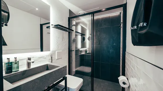 Modern and clean hotel bathroom with a sleek sink, illuminated mirror, toilet, and a glass-enclosed shower. A hair dryer bag hangs on the wall.