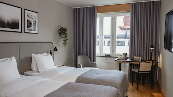 To pent oppreide senger med grå sengedekor står i et moderne hotellrom. Et stort vindu med lilla gardiner, et lite skrivebord og veggkunst fullfører den koselige innstillingen.